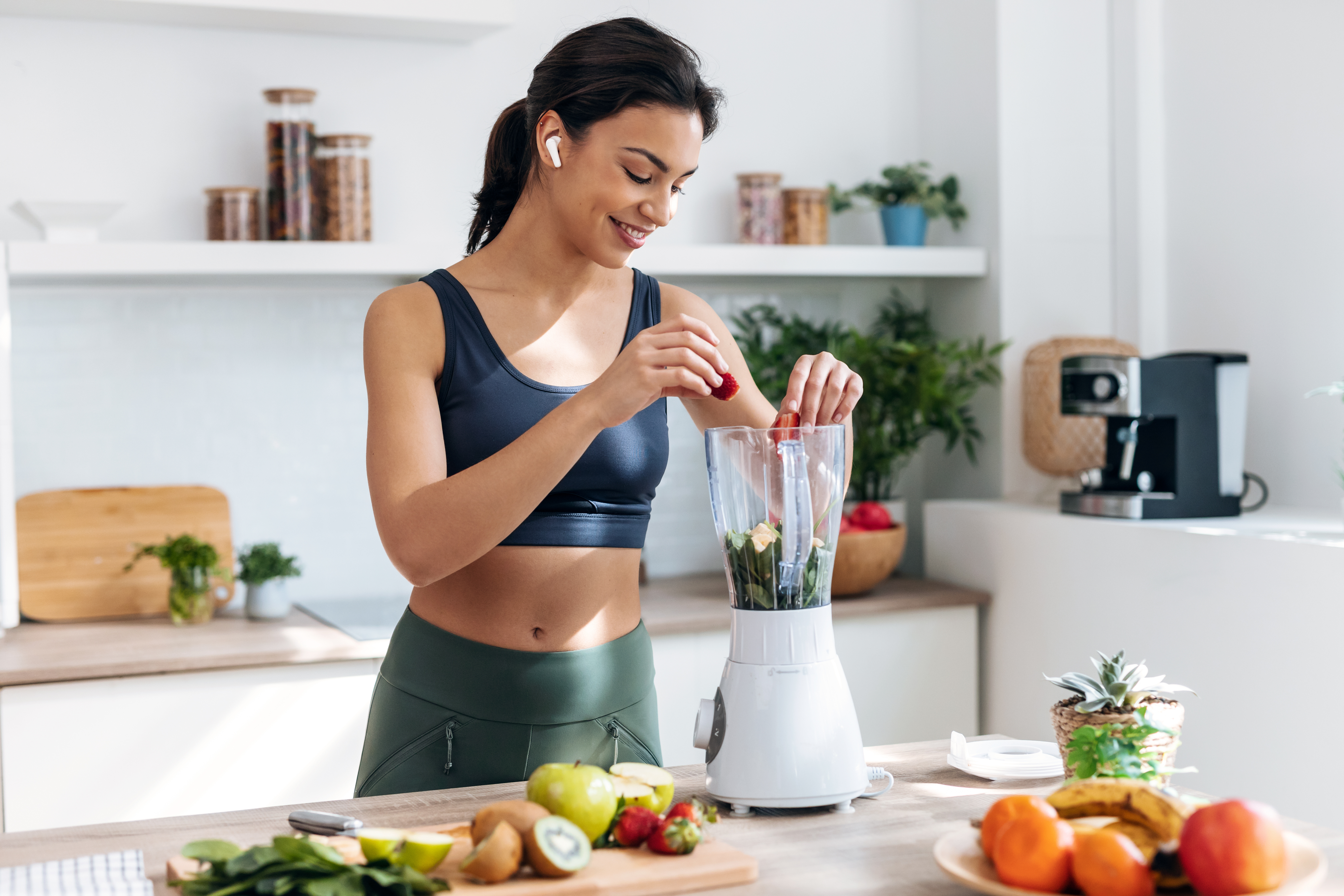 Shot of athletic woman preparing smothie with vegetables and fruits while listening music with earphones in the kitchen at home.